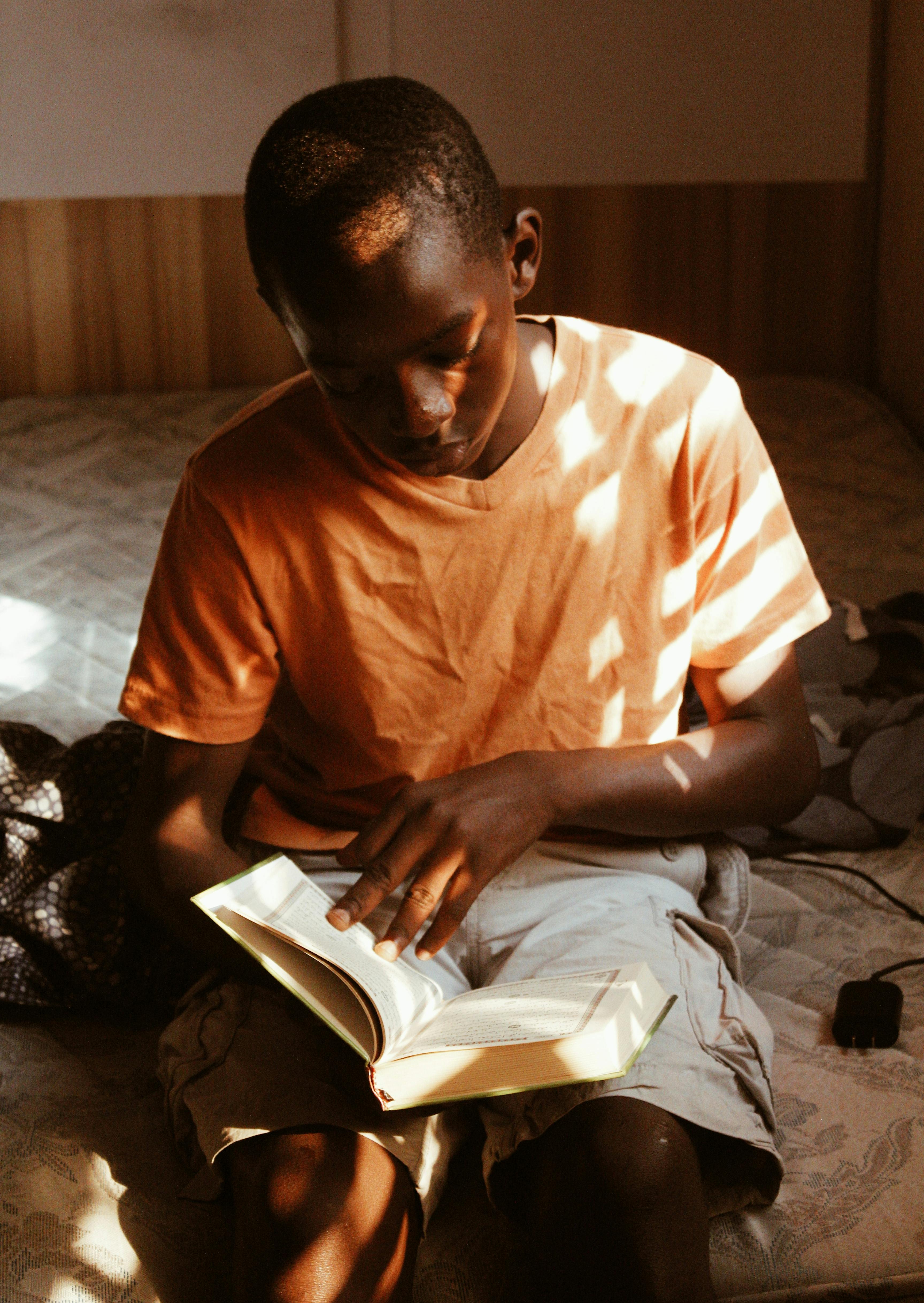 A young boy reading a book in a sunlit room with light and shadow play.